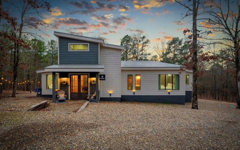 Modern house with a gravel yard, surrounded by trees, under a colorful sunset sky, with warm lights inside and outside the house.